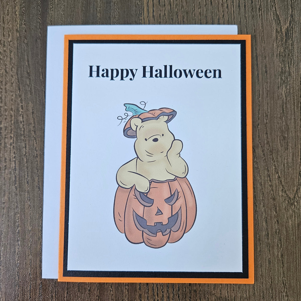 A handmade Halloween greeting card featuring Winnie the Pooh holding a carved pumpkin, with the text 'Happy Halloween' at the top. The card has an orange border, a black inner border, and is photographed standing on a dark wooden surface against a light gray wall.
