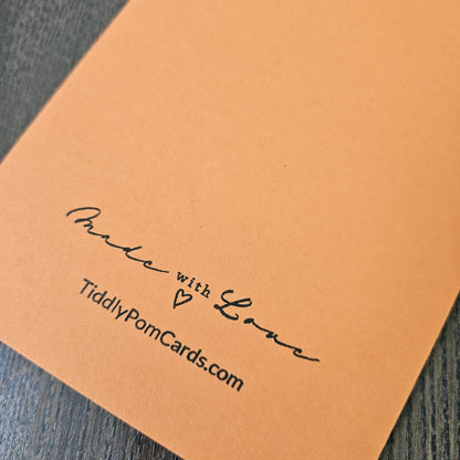 A close-up of a peach-colored paper card with black handwritten text reading 'Made with Love' and 'TiddlyPomCards.com', placed on a dark wood surface.