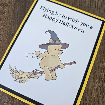 A square Halloween greeting card featuring a cartoon teddy bear wearing a witch hat and flying on a broomstick, with the text 'Flying by to wish you a Happy Halloween.' The card has a white center, a yellow border, and a black inner border, photographed on a brown wood surface.