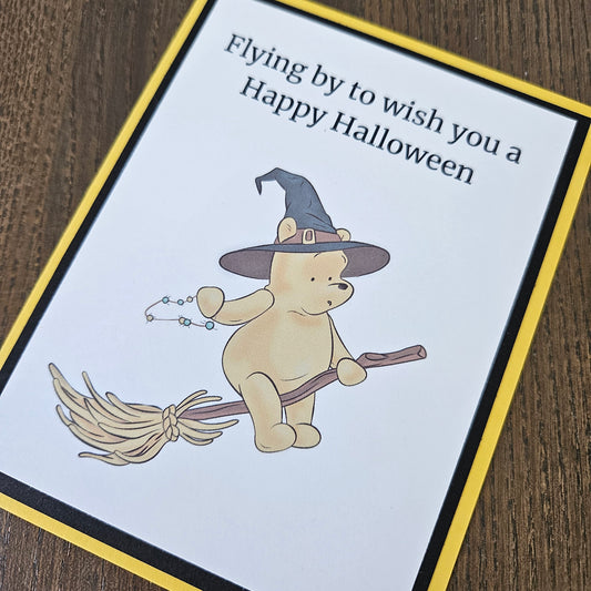 A square Halloween greeting card featuring a cartoon teddy bear wearing a witch hat and flying on a broomstick, with the text 'Flying by to wish you a Happy Halloween.' The card has a white center, a yellow border, and a black inner border, photographed on a brown wood surface.