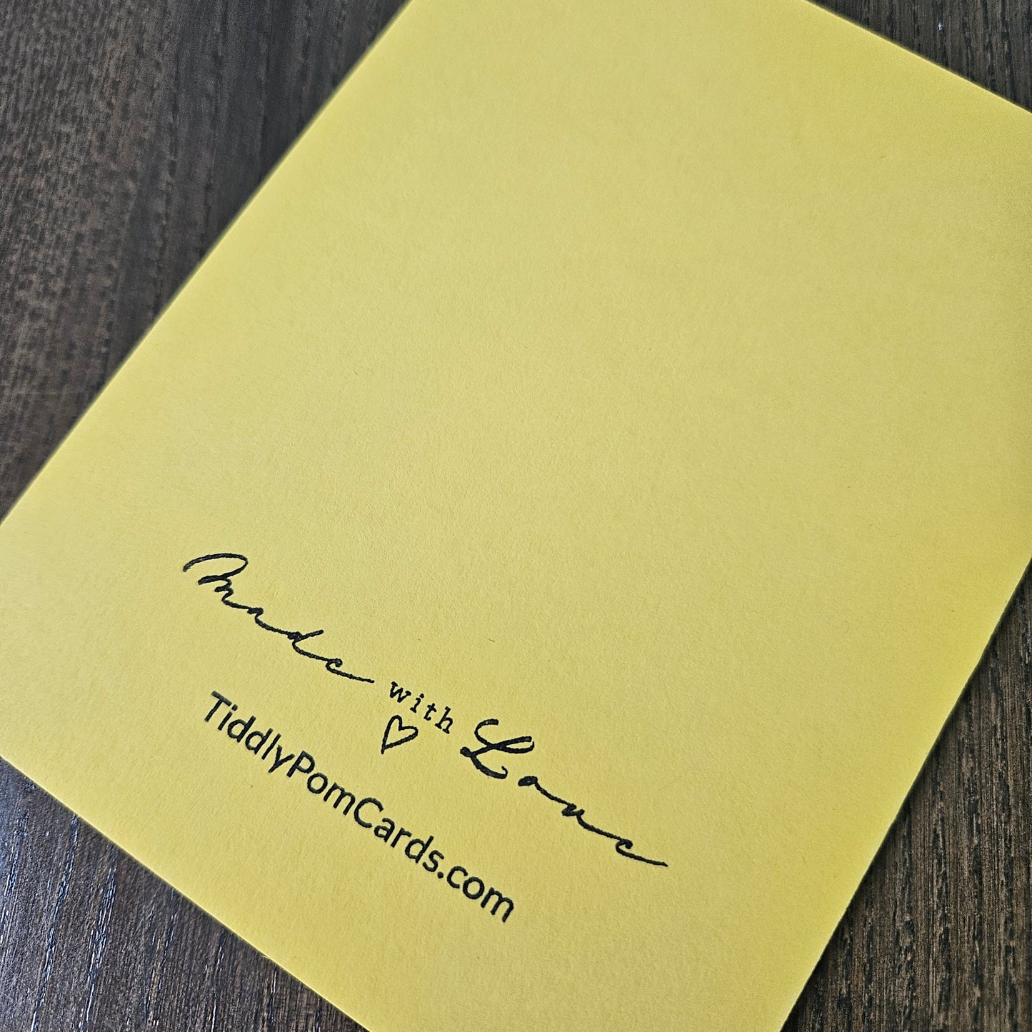 A square pale yellow greeting card with black printed text reading 'Made with Love' and 'TidilyPomCards.com', photographed on a dark wood surface.