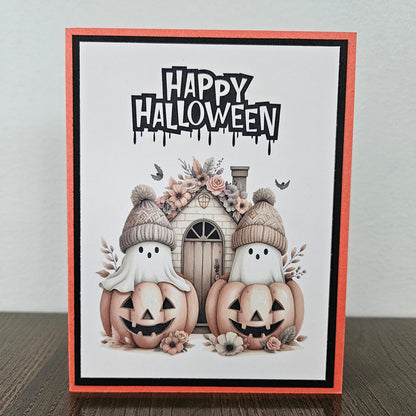 Front of a handmade A2 cute boho ghost Halloween card featuring layered orange and black cardstock framing two ghosts wearing beanie hats inside jack-o'-lantern pumpkins with text reading HAPPY HALLOWEEN.