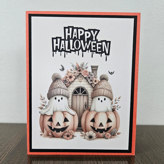 Front of a handmade A2 cute boho ghost Halloween card featuring layered orange and black cardstock framing two ghosts wearing beanie hats inside jack-o'-lantern pumpkins with text reading HAPPY HALLOWEEN.