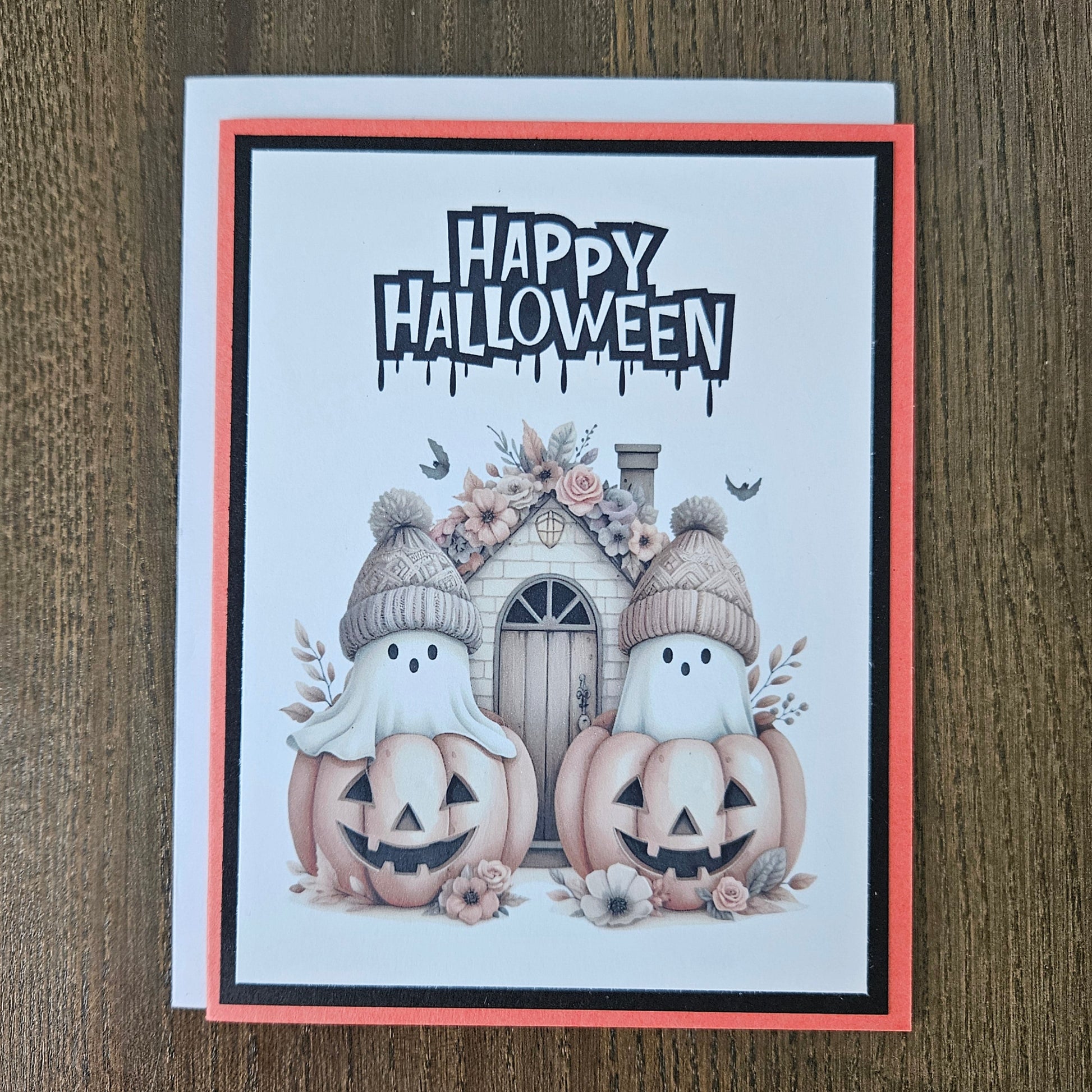 Front of a handmade A2 cute boho ghost Halloween card featuring layered orange and black cardstock framing two ghosts wearing beanie hats inside jack-o'-lantern pumpkins with text reading HAPPY HALLOWEEN.