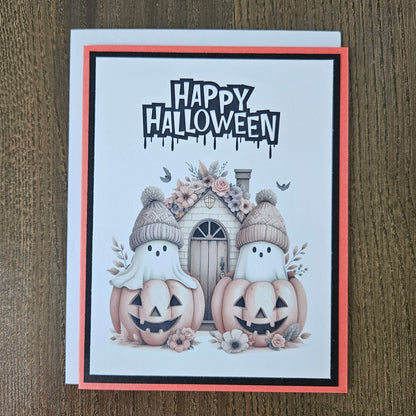 Front of a handmade A2 cute boho ghost Halloween card featuring layered orange and black cardstock framing two ghosts wearing beanie hats inside jack-o'-lantern pumpkins with text reading HAPPY HALLOWEEN.