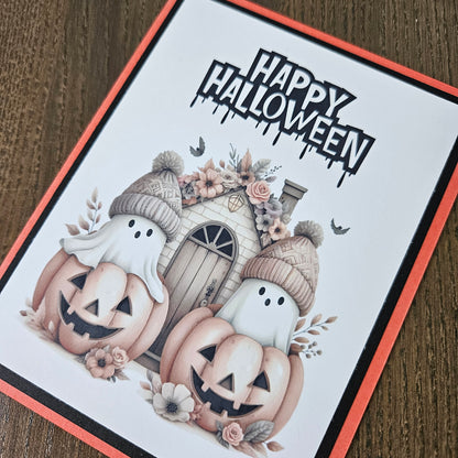 Front of a handmade A2 cute boho ghost Halloween card featuring layered orange and black cardstock framing two ghosts wearing beanie hats inside jack-o'-lantern pumpkins with text reading HAPPY HALLOWEEN.