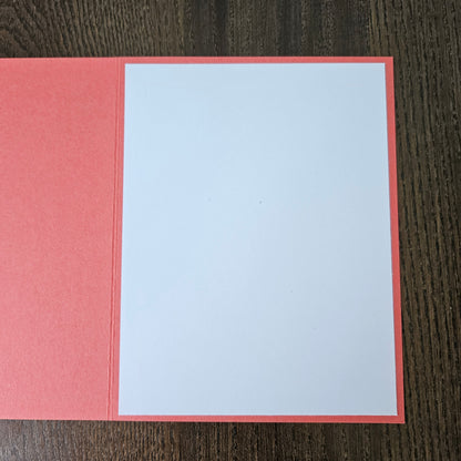 A blank greeting card with a coral pink border and matching envelope on a dark wood background.