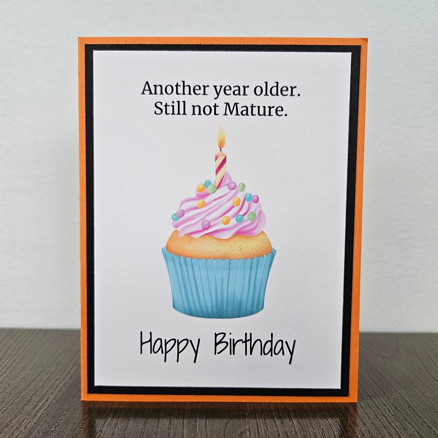 A handmade birthday card with an orange border and black inner frame, standing on a dark wood surface against a light gray wall. The card features a cupcake illustration with pink frosting, a blue wrapper, and a single candle, with the text 'Another year older.