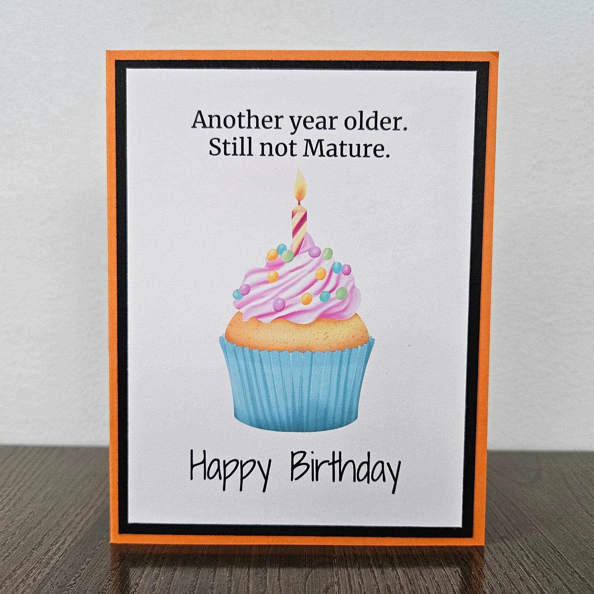 A handmade birthday card with an orange border and black inner frame, standing on a dark wood surface against a light gray wall. The card features a cupcake illustration with pink frosting, a blue wrapper, and a single candle, with the text 'Another year older.