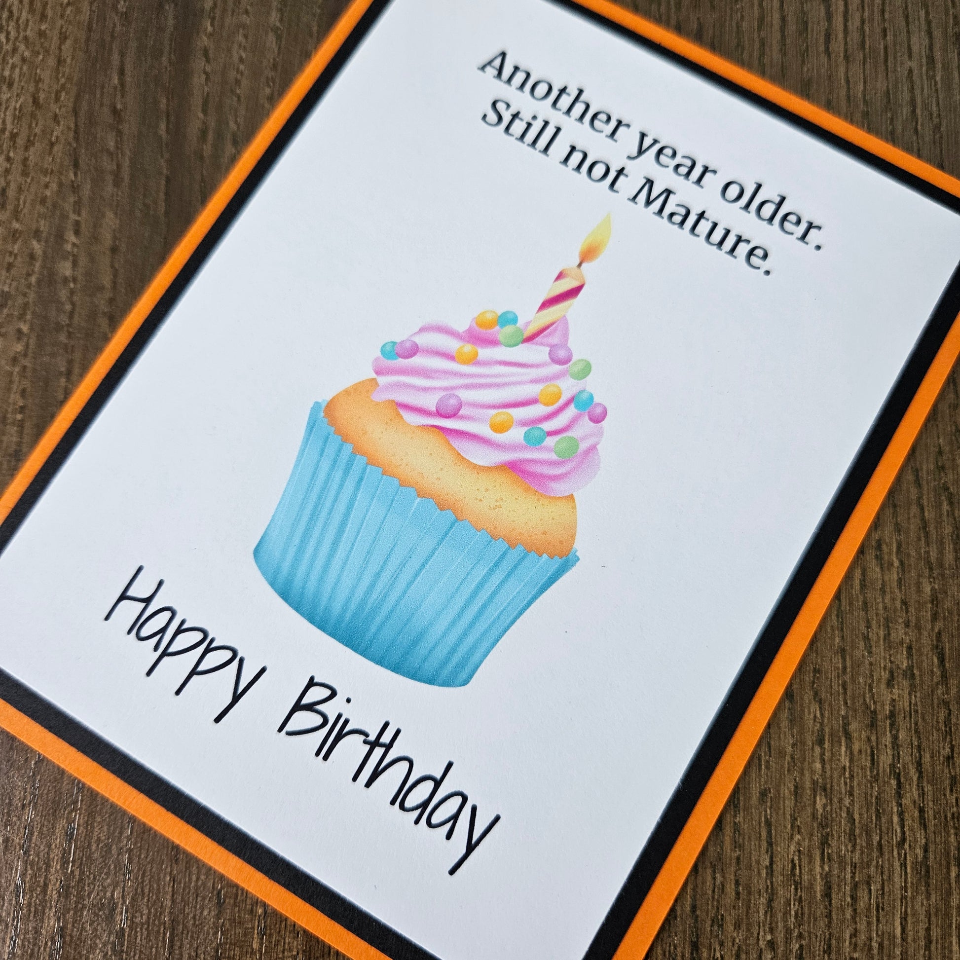 A square birthday card with an orange border and black inner border, placed on a wooden surface. The card features a watercolor-style cupcake with pink frosting, colorful sprinkles, and a candle, along with the text 'Another year older.