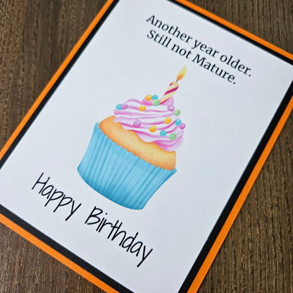 A square birthday card with an orange border and black inner border, placed on a wooden surface. The card features a watercolor-style cupcake with pink frosting, colorful sprinkles, and a candle, along with the text 'Another year older.