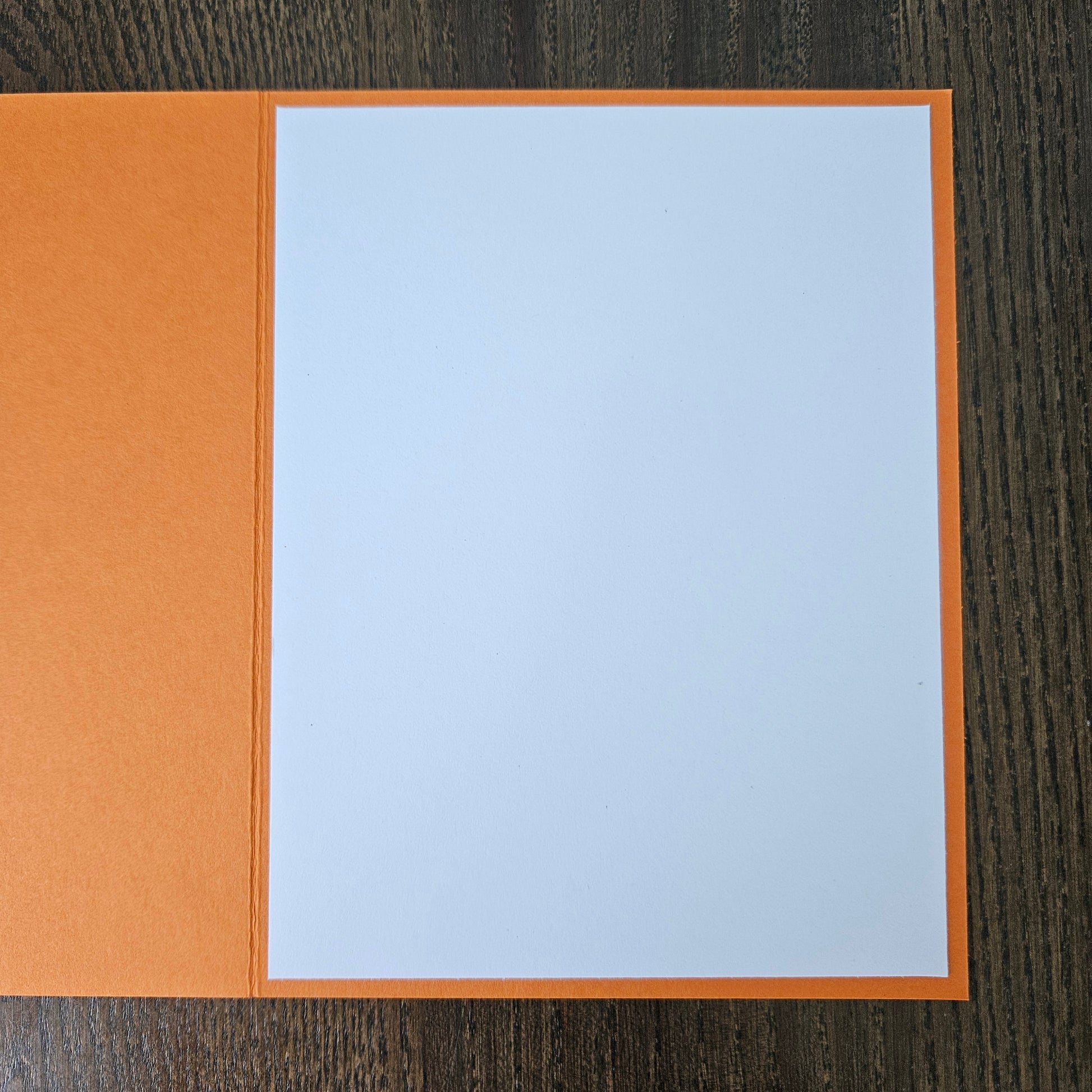 An open greeting card with a textured orange cover and a blank white center panel, photographed from above on a dark wood surface.