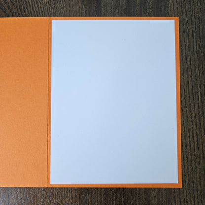 An open greeting card with a textured orange cover and a blank white center panel, photographed from above on a dark wood surface.