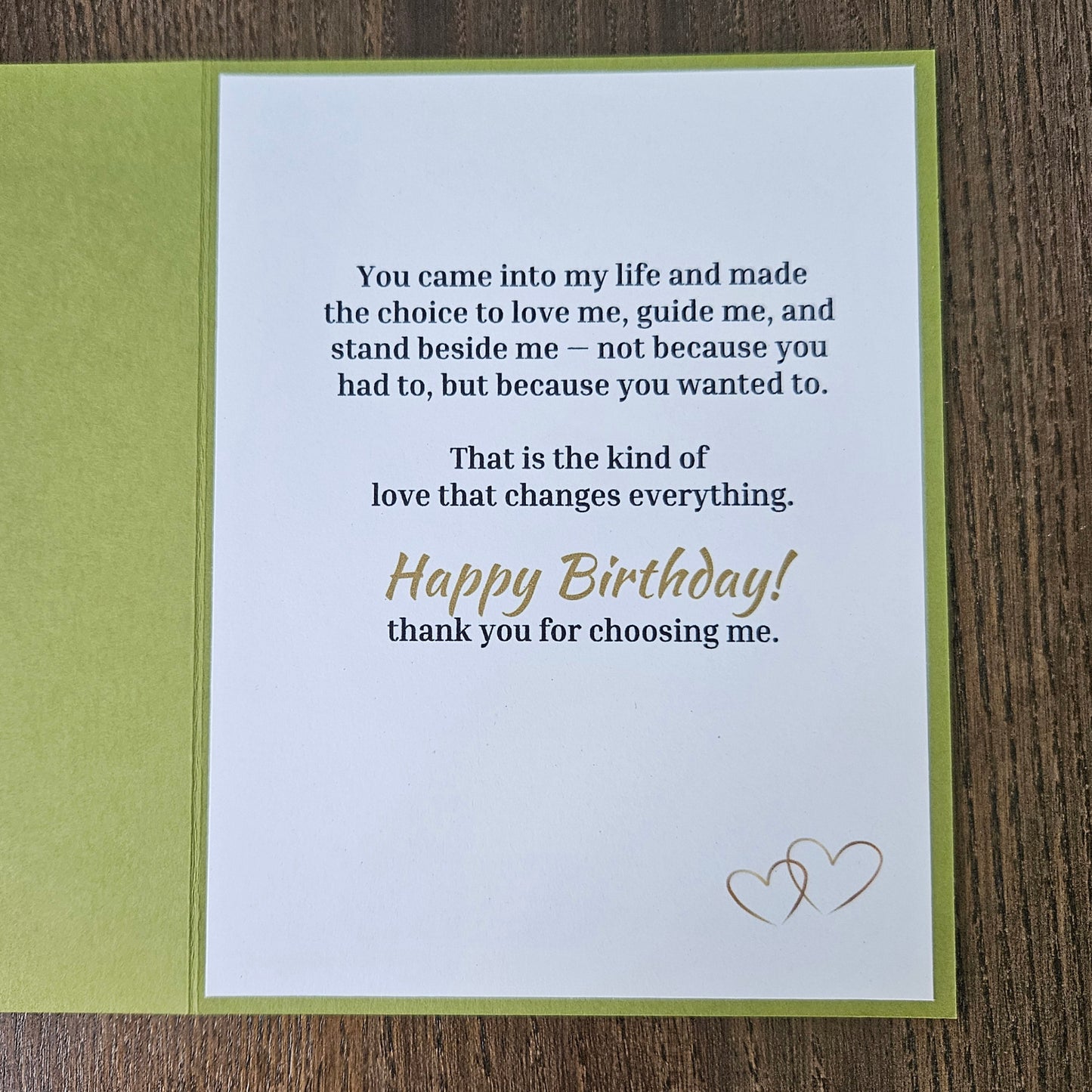 A square birthday card with a white front insert and olive green backing, placed on a brown wood surface. The card features a sentimental printed message in black text and 'Happy Birthday' in gold script, with a small gold heart design in the upper right.