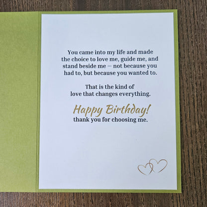 A square birthday card with a white front insert and olive green backing, placed on a brown wood surface. The card features a sentimental printed message in black text and 'Happy Birthday' in gold script, with a small gold heart design in the upper right.
