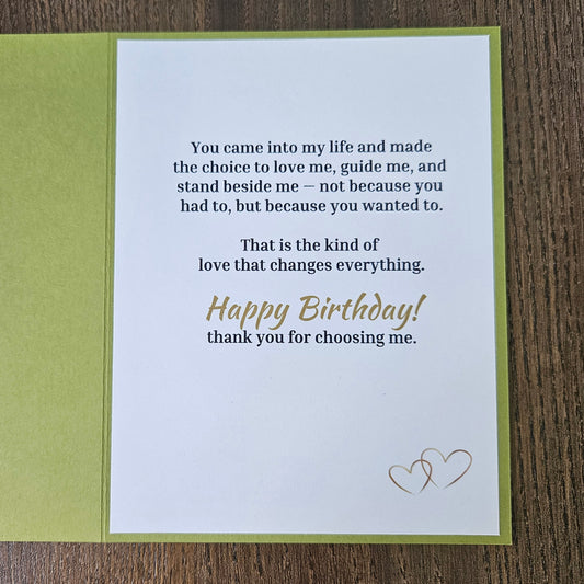 A square birthday card with a white front insert and olive green backing, placed on a brown wood surface. The card features a sentimental printed message in black text and 'Happy Birthday' in gold script, with a small gold heart design in the upper right.