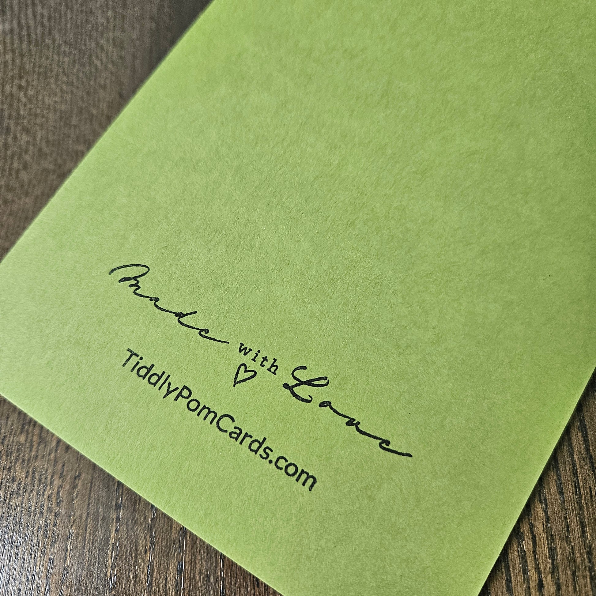 A close-up of a light olive green pom card printed with 'Made with Love' and 'TiddlyPomCards.com' in black ink, resting on a dark wood surface.