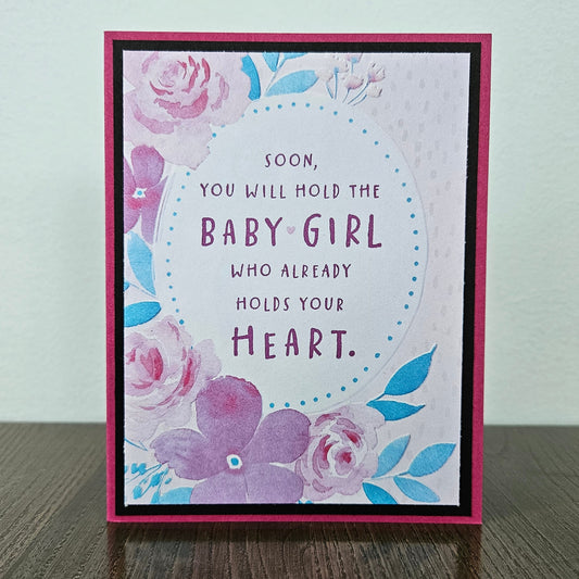 Front of a handmade A2 new baby girl card with a mauve border framing a watercolor floral oval design with text reading Soon you will hold the Baby Girl who already holds your Heart.