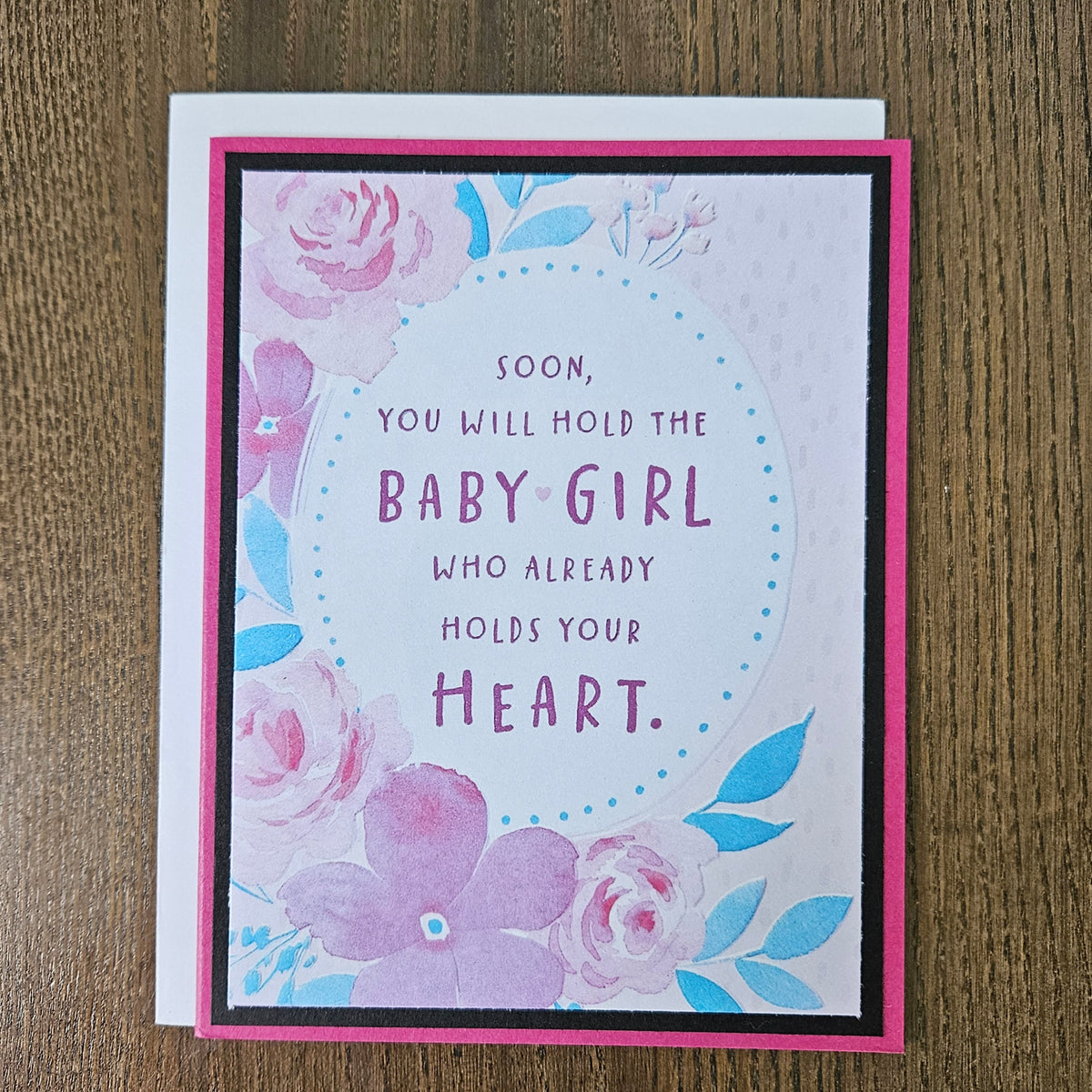 Front of a handmade A2 new baby girl card with a mauve border framing a watercolor floral oval design with text reading Soon you will hold the Baby Girl who already holds your Heart.