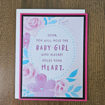 Front of a handmade A2 new baby girl card with a mauve border framing a watercolor floral oval design with text reading Soon you will hold the Baby Girl who already holds your Heart.