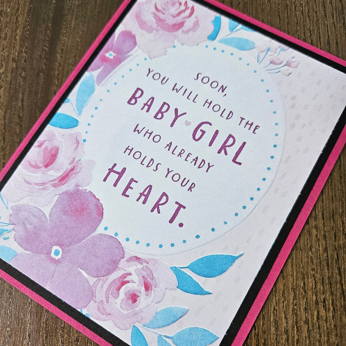 A close-up of a handmade greeting card featuring a pink border, watercolor floral designs in pink and blue, and the printed message 'SOON YOU WILL HOLD THE BABY GIRL WHO ALREADY HOLDS YOUR HEART.' on a wooden surface.