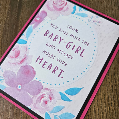 A close-up of a handmade greeting card featuring a pink border, watercolor floral designs in pink and blue, and the printed message 'SOON YOU WILL HOLD THE BABY GIRL WHO ALREADY HOLDS YOUR HEART.' on a wooden surface.