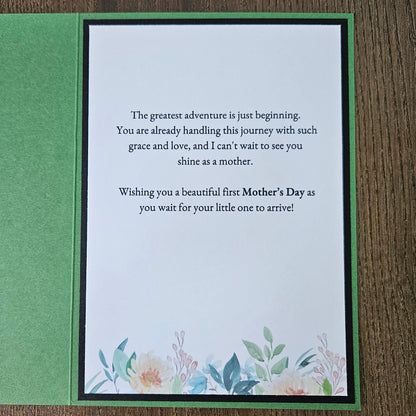 A square Mother's Day greeting card with a green border and a white center panel featuring a floral quote and watercolor flowers, placed on a wooden surface.