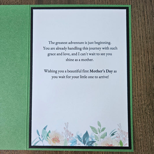 A square Mother's Day greeting card with a green border and a white center panel featuring a floral quote and watercolor flowers, placed on a wooden surface.