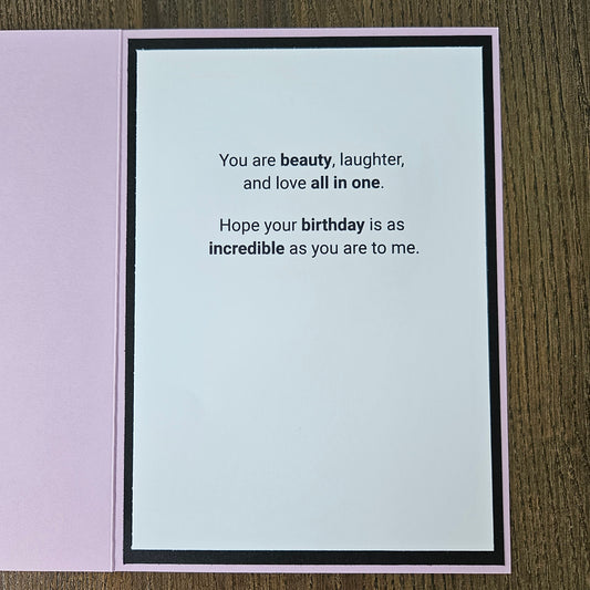 An open square greeting card with a light pink border and a white center panel printed with a birthday message in black text, placed on a wood surface.