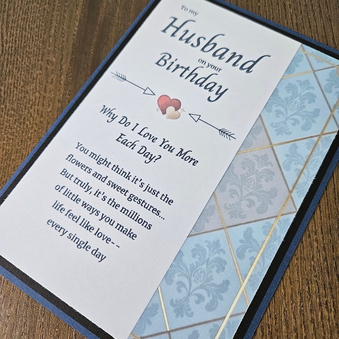 A close-up of a printed card or bookmark-style gift item placed on a wooden surface. The visible text reads 'Husband on your Birthday' with decorative arrows, a small pink heart, and a quote about love.