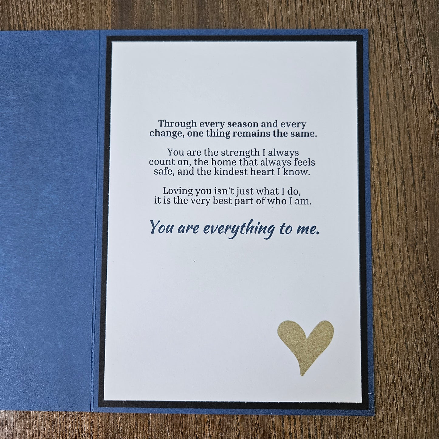A square blue greeting card or gift card shown open on a wooden surface, with a white front insert featuring a gold heart and printed sentimental text, plus a blue backing piece.