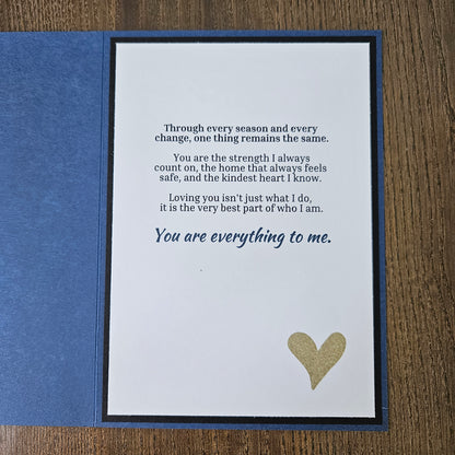 A square blue greeting card or gift card shown open on a wooden surface, with a white front insert featuring a gold heart and printed sentimental text, plus a blue backing piece.