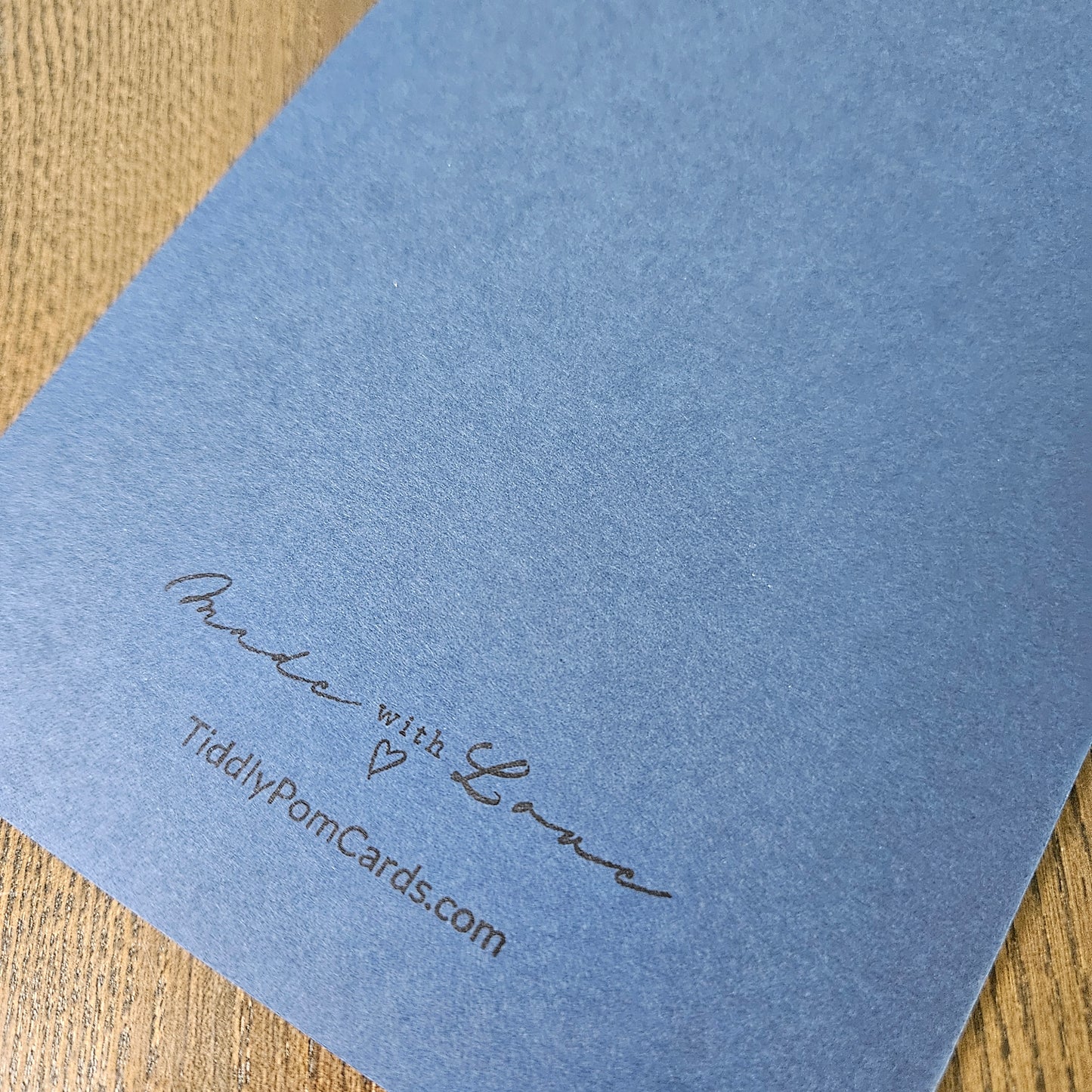 A close-up of a light blue textured card with the words 'mix with Love' and 'TiddlyPomCards.com' printed in black script on a wooden surface.