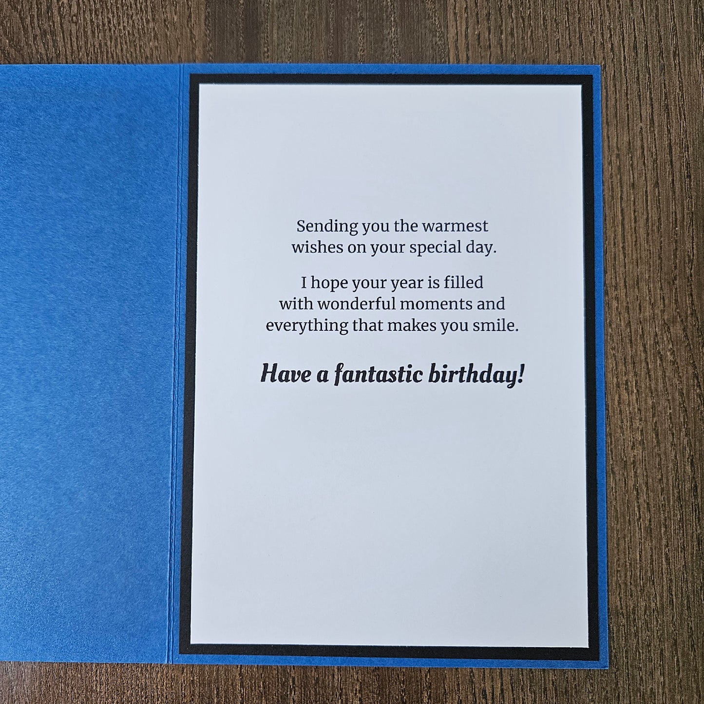 A square blue greeting card or notebook cover shown open on a wood surface, with a white insert featuring black text that reads, 'Sending you the warmest birthday wishes on your special day. I hope your year is filled with wonderful moments and everything that makes you smile.