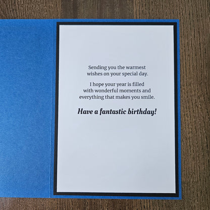 A square blue greeting card or notebook cover shown open on a wood surface, with a white insert featuring black text that reads, 'Sending you the warmest birthday wishes on your special day. I hope your year is filled with wonderful moments and everything that makes you smile.