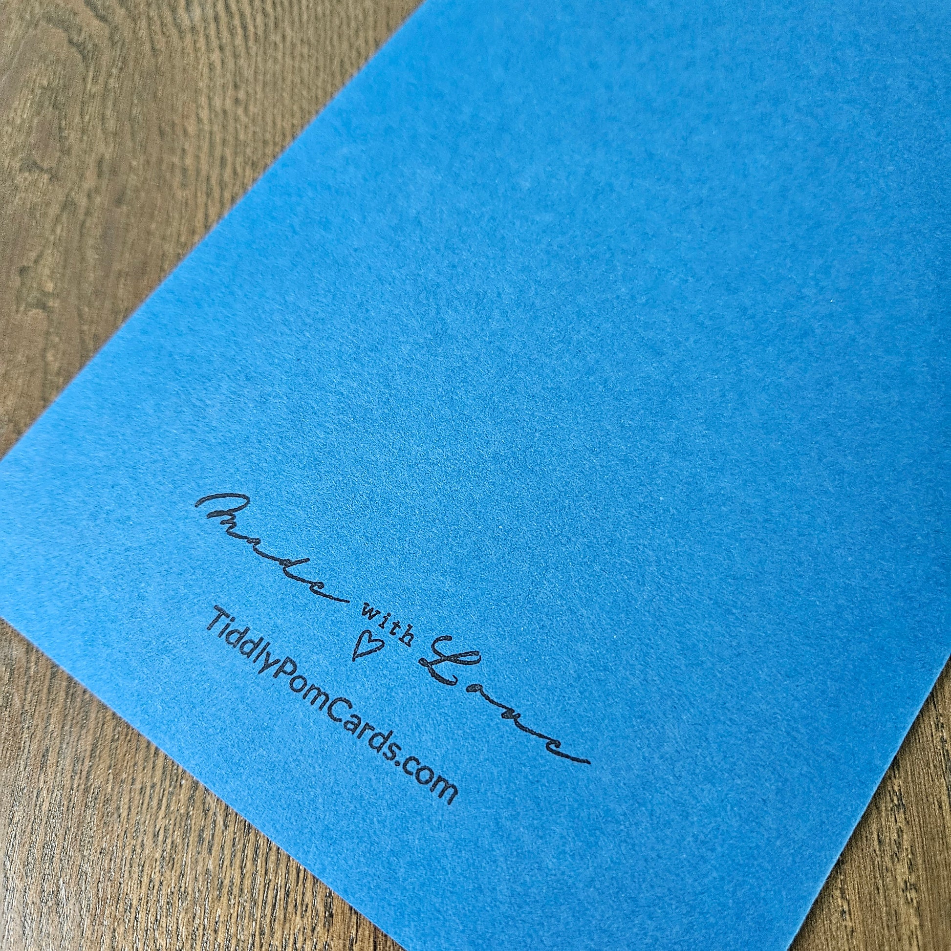A close-up of a blue textured card or paper sheet placed on a wooden surface, with black printed text reading 'Make with Love' and 'TiddlyPonkCards.com' in the lower right corner.