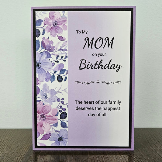 A handmade greeting card with a lavender border, featuring a floral watercolor pattern in purple and pink on the left side and a message for 'MOM' on the right, displayed on a dark wooden surface against a light gray wall.