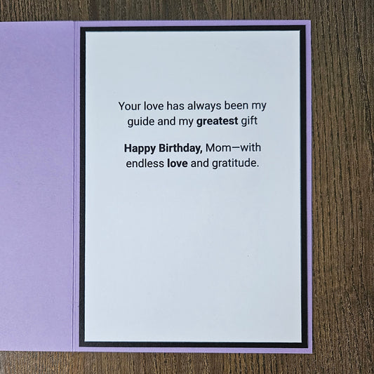 A square birthday card with a lavender purple border and a white center panel printed with the message, 'Your love has always been my guide and my greatest gift Happy Birthday, Mom—with endless love and gratitude.' The card is photographed flat on a brown wood-grain surface.