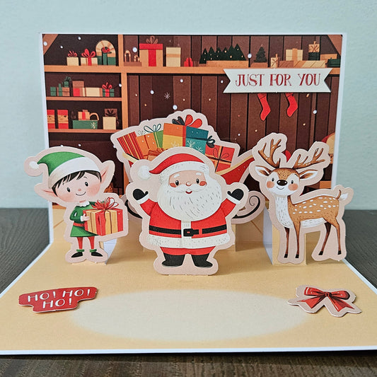 Front of a handmade 5x7 3D pop-up Christmas card featuring Santa Claus, an elf, and a reindeer surrounded by presents with text reading Merry Christmas.