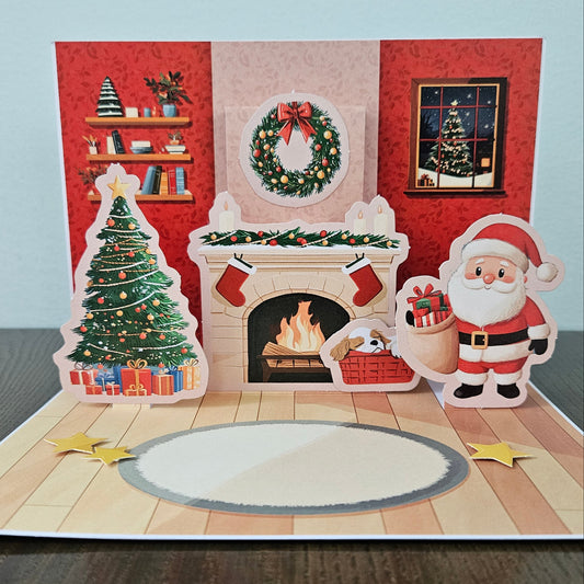 A 3D pop-up Christmas greeting card displayed on a dark wooden table against a light blue-gray wall. The card features a festive holiday scene with a decorated Christmas tree, a fireplace with a wreath and burning fire, a window showing a snowy tree, and a cartoon Santa Claus holding a sack of gifts.