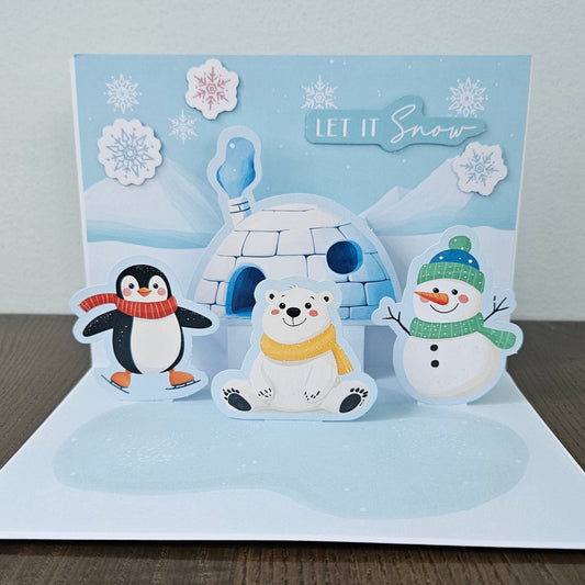 Front of a handmade 5x7 3D pop-up Christmas card featuring an igloo, snowman, penguin, and polar bear with text reading Happy Holidays.