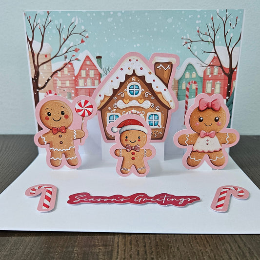 Front of a handmade 5x7 3D pop-up Christmas card featuring a gingerbread house and family with candy cane accents and text reading With Love.