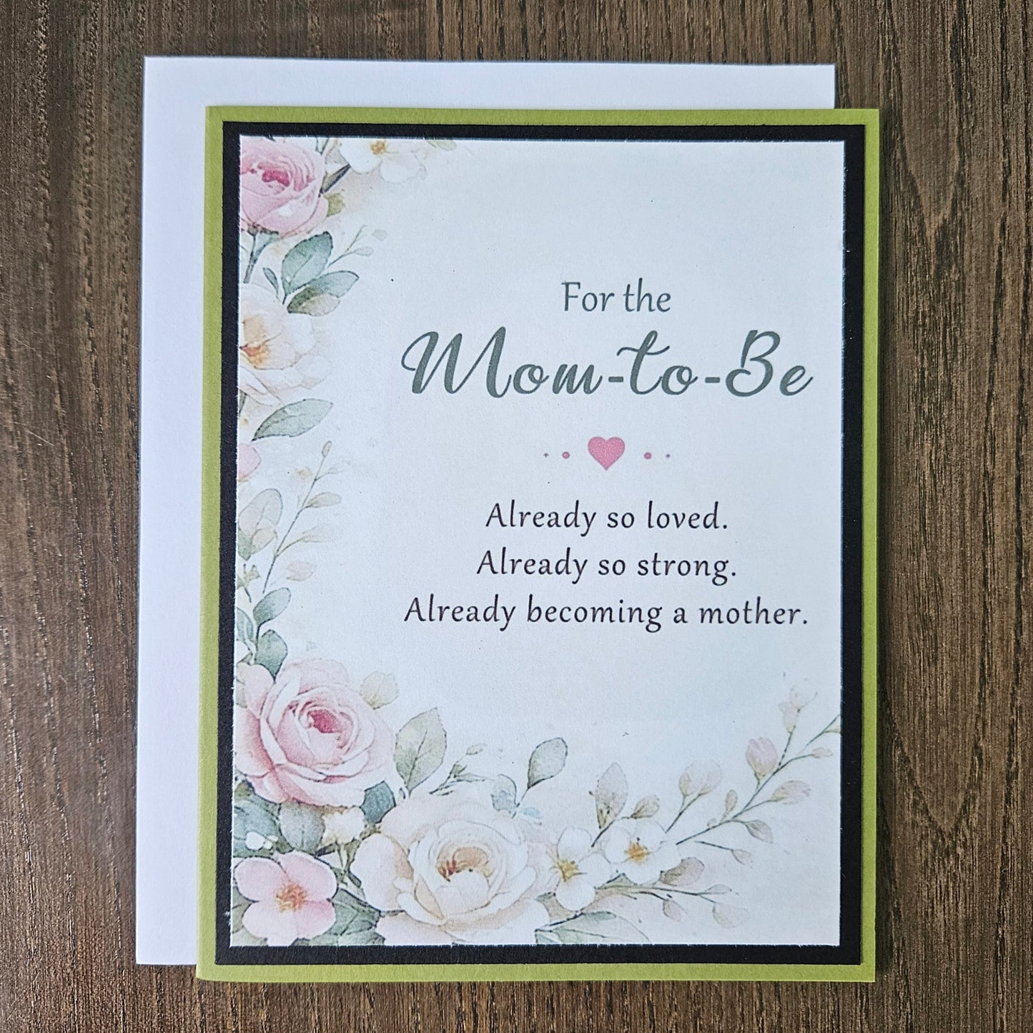 A floral 'Mom-to-Be' greeting card with a white envelope, displayed flat on a dark wood background. The card features watercolor-style pink and white roses with green leaves, bordered by black and olive green stripes.
