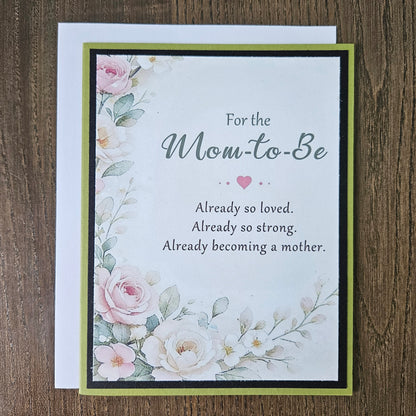 A floral 'Mom-to-Be' greeting card with a white envelope, displayed flat on a dark wood background. The card features watercolor-style pink and white roses with green leaves, bordered by black and olive green stripes.