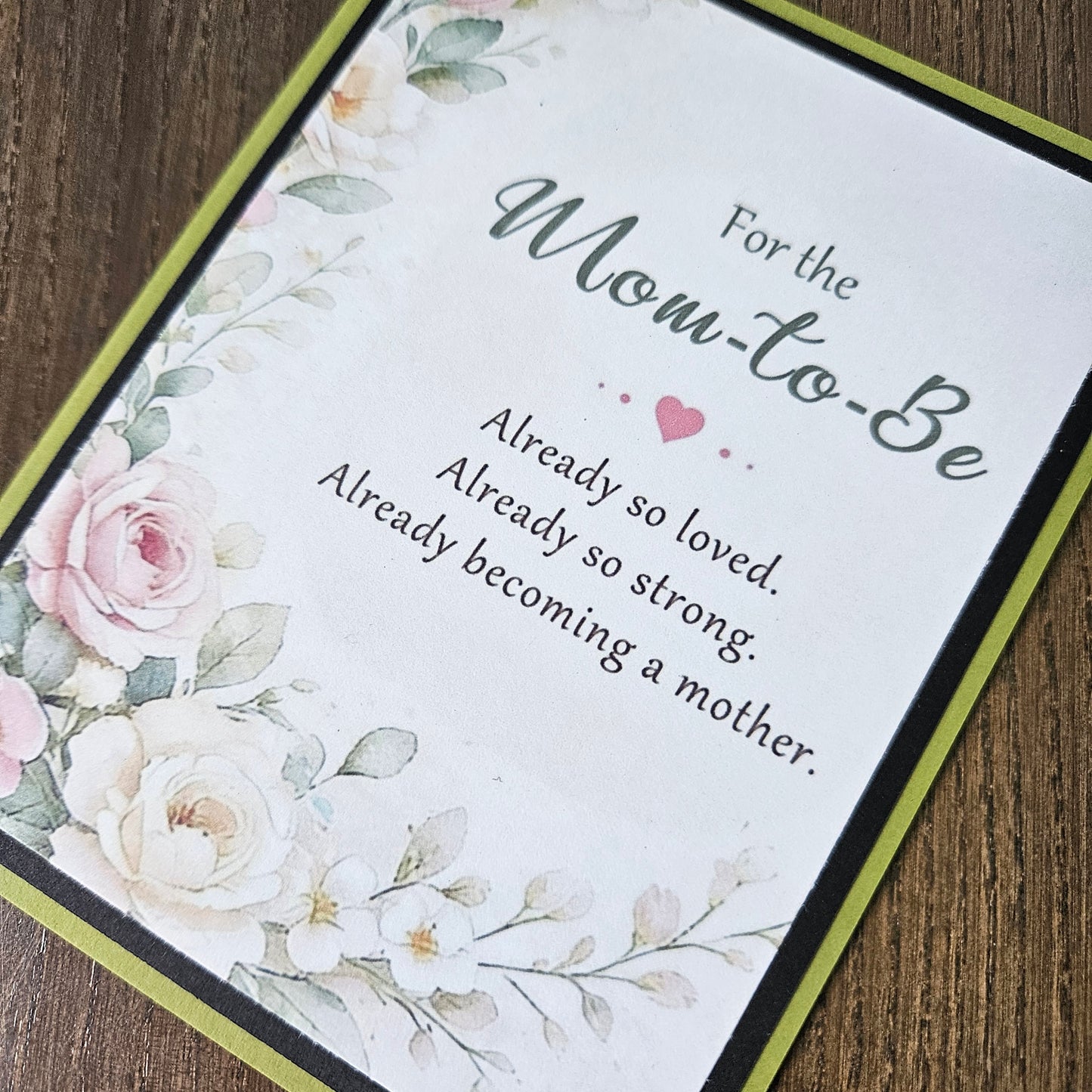 A square floral greeting card for a 'Mom-to-Be' laid diagonally on a dark wood background. The card features a white background with watercolor-style pink and white roses, green leaves, and elegant gray text that reads 'For the Mom-to-Be Already so loved.