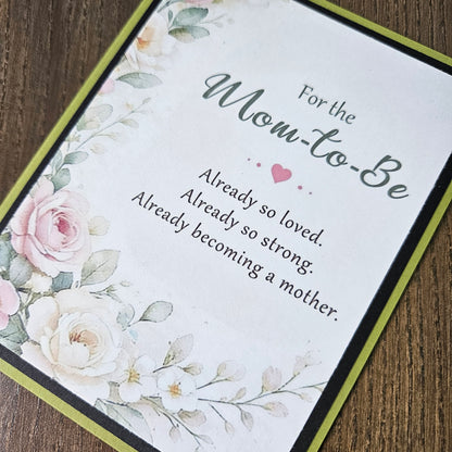 A square floral greeting card for a 'Mom-to-Be' laid diagonally on a dark wood background. The card features a white background with watercolor-style pink and white roses, green leaves, and elegant gray text that reads 'For the Mom-to-Be Already so loved.