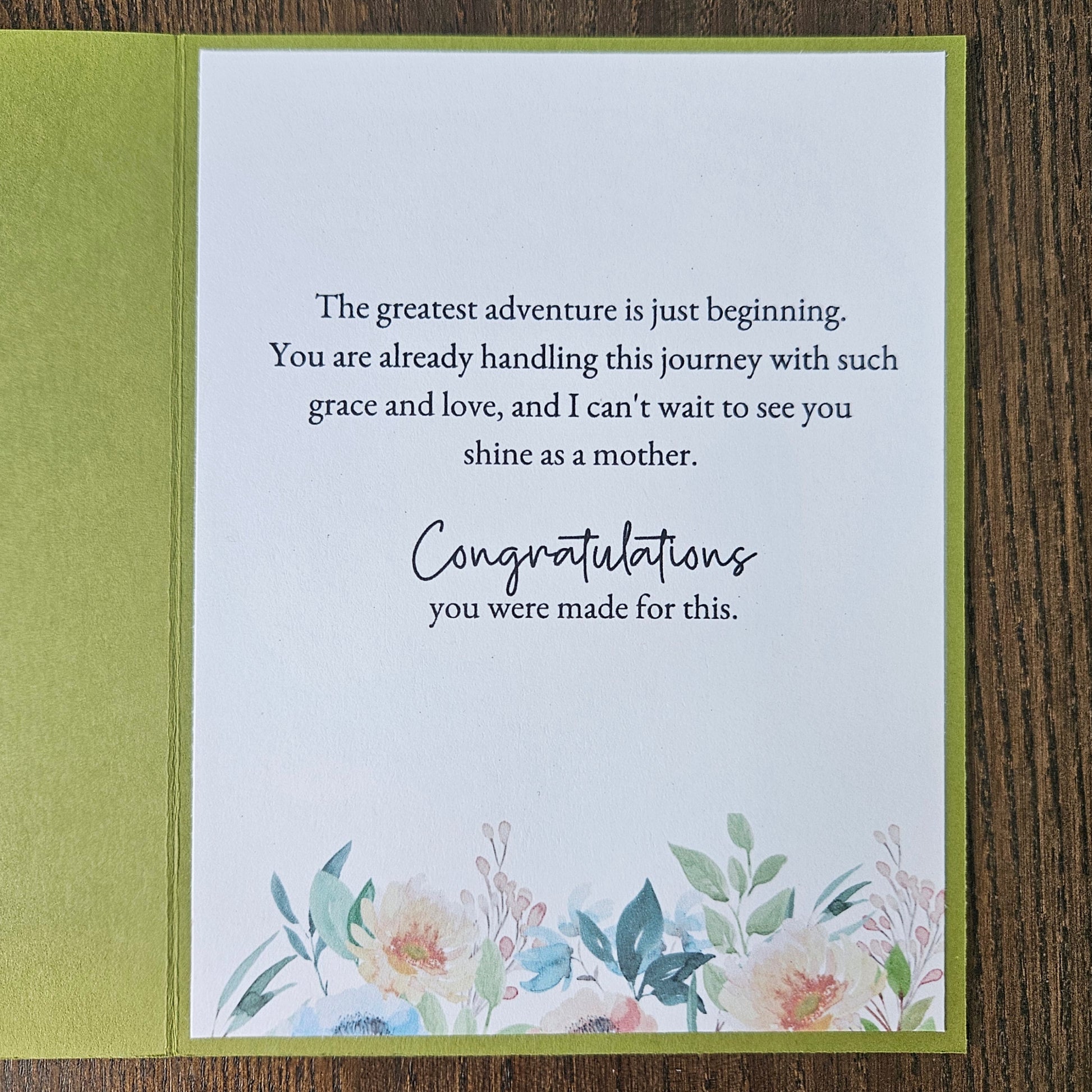 An open greeting card with a white front insert featuring a motivational quote and watercolor floral illustration at the bottom, set against an olive green backing on a dark wood surface.