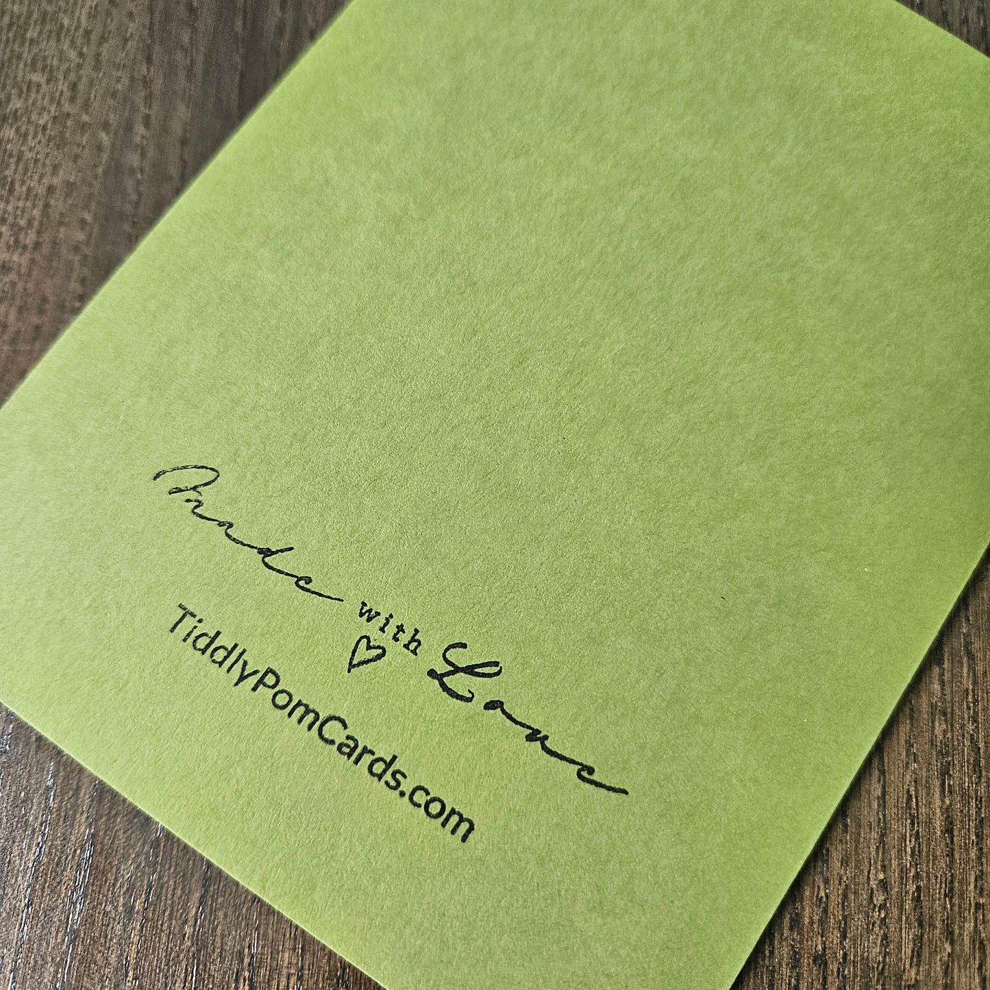 A square olive green card with black printed text reading 'Make with Love' and 'TiddlyPomCards.com' placed on a dark wood surface.