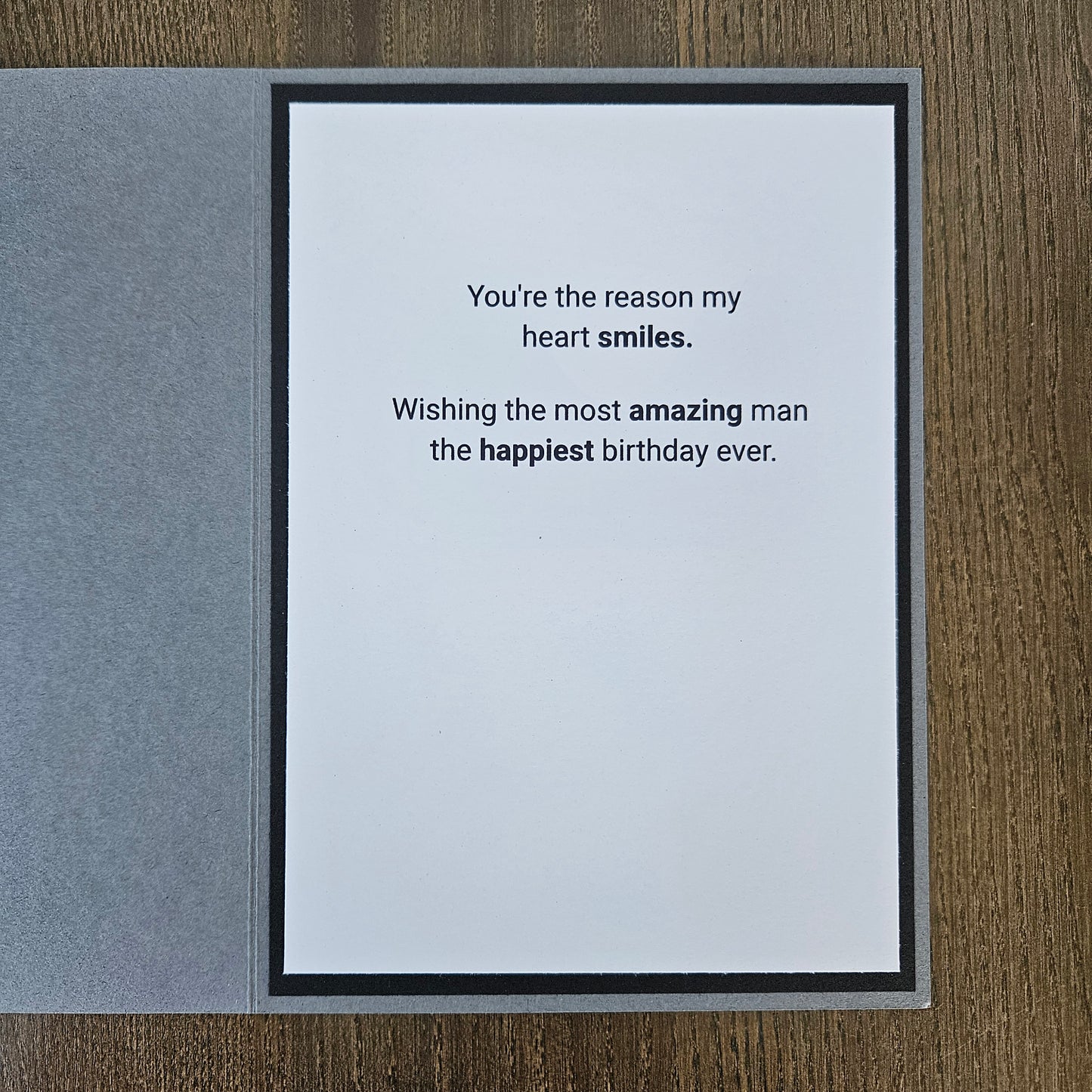 A square greeting card with a white center panel and black border, placed on a gray textured cover, photographed on a wood surface. The card reads: 'You're the reason my heart smiles.