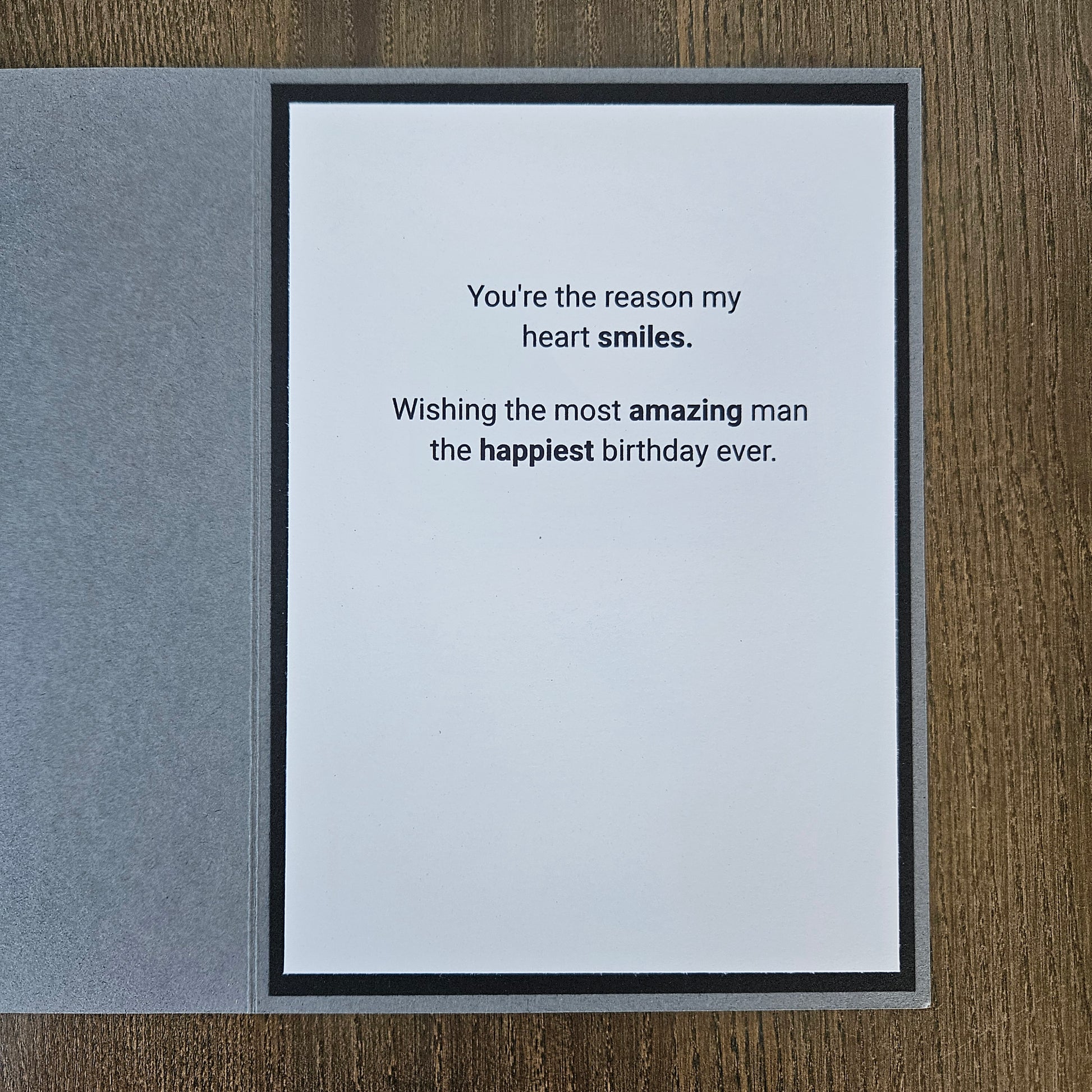 A square greeting card with a white center panel and black border, placed on a gray textured cover, photographed on a wood surface. The card reads: 'You're the reason my heart smiles.
